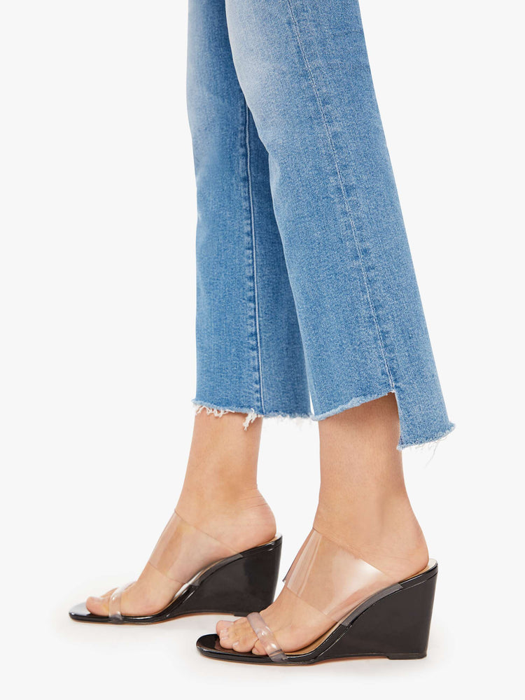 Hem close up view of a woman in a mid blue high-waisted bootcut is cropped at the ankle with a frayed step-hem.