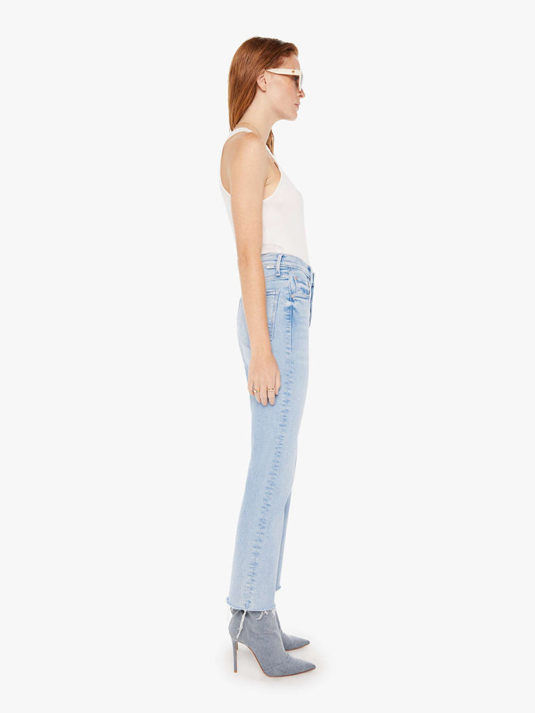 Side view of a woman wearing a light blue wash jean featuring a high rise, an ankle length flare with frayed hem, paired with a white tank top.