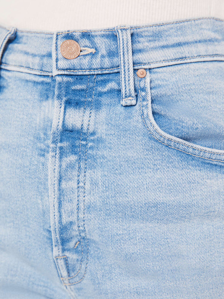 A close up swatch detail view of a light blue wash jean featuring light gold hardware and contrast beige stitching.