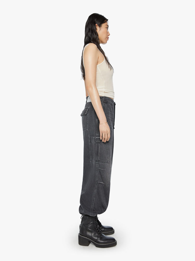 Side view of a woman super high-waisted wide-leg jeans feature a drawstring waist, oversized utility pockets and an ankle-length inseam with an elastic drawstring at the hem in a faded black wash.
