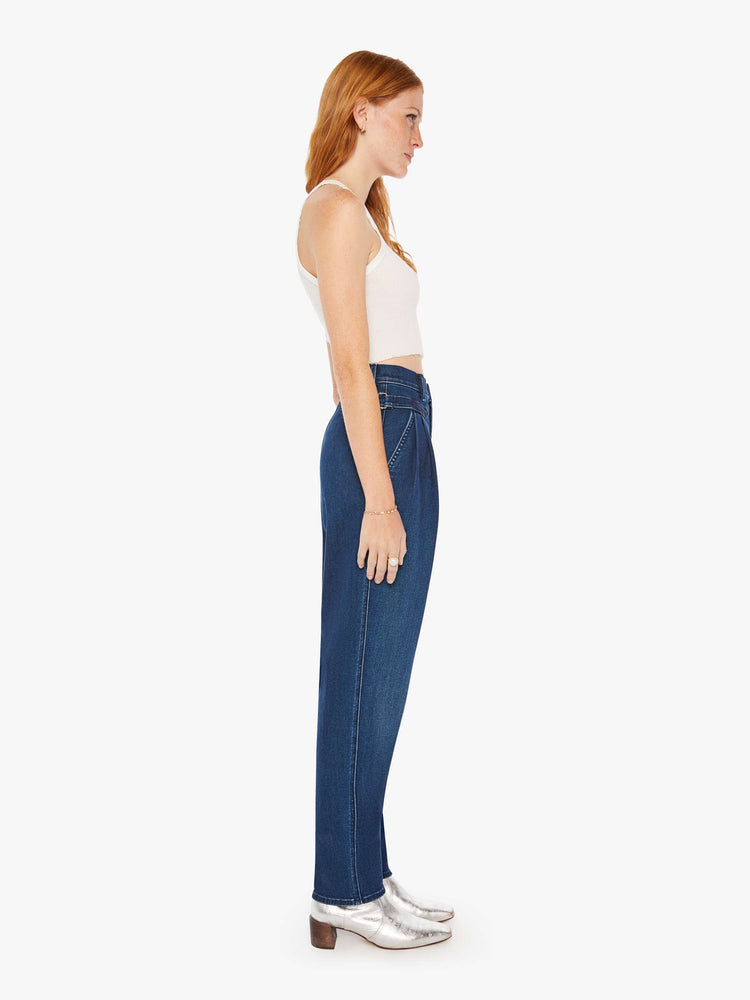 Side view of a woman super high-rise jeans with pleated, adjustable tabs below the waist, side slit pockets, a long 31-inch inseam and a tapered straight leg in a dark blue wash.