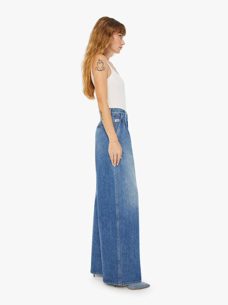 Side view of a woman medium blue super high-rise, wide-leg jeans are designed with a pleated waistband, slightly dropped crotch, side slit pockets and a long inseam.
