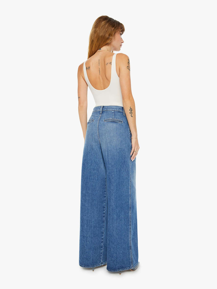 Back view of a woman medium blue super high-rise, wide-leg jeans are designed with a pleated waistband, slightly dropped crotch, side slit pockets and a long inseam.