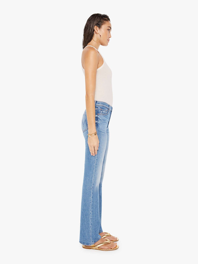 Side full body view of a woman wearing a medium blue wash jean featuring a mid rise and a full length flare inseam.