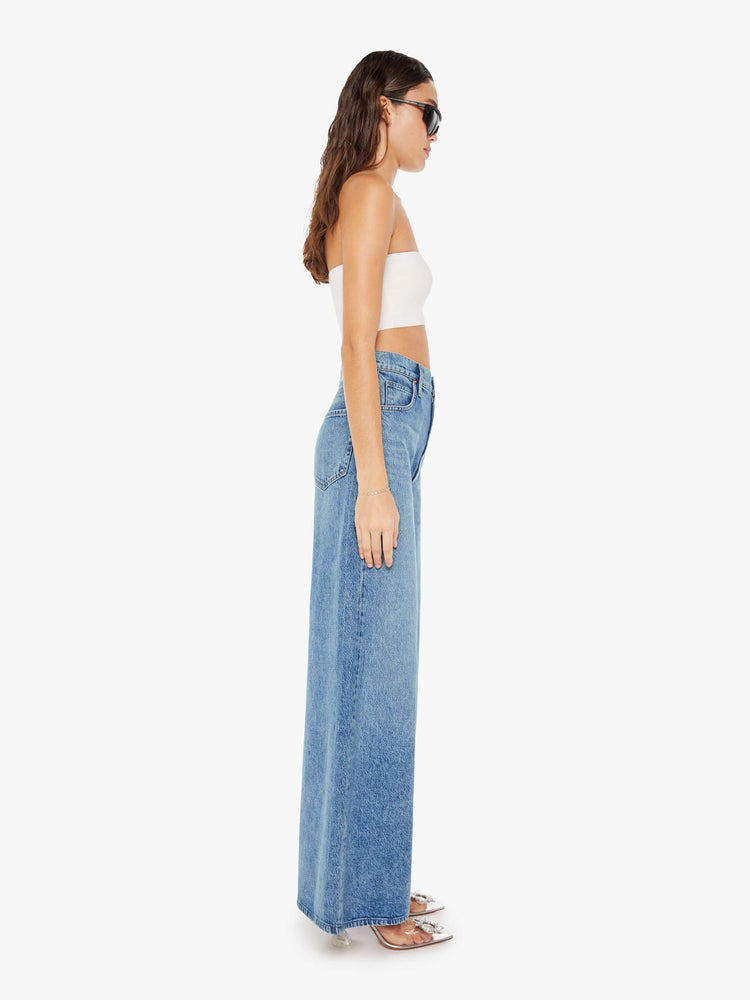 Side view of a woman wearing a medium blue wash jean featuring a super high rise and a wide long leg.