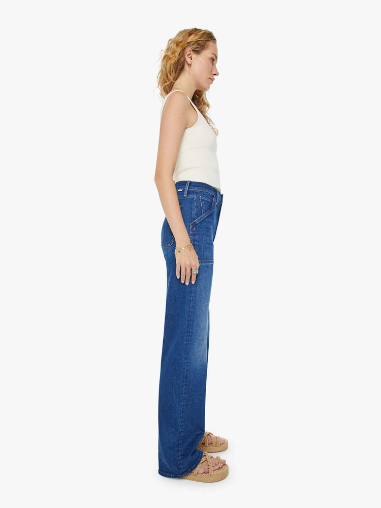 Side view of a woman in dark blue super high-waisted jeans with a loose wide leg, utility-inspired patch pockets, and subltle whiskering and fading at the knee. Styled with a white tank top.