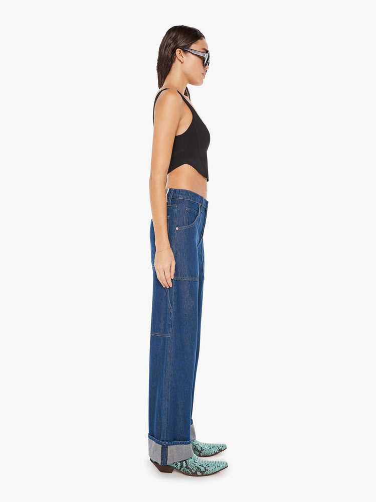 Side view of a womens dark blue denim jean featuring a long cuffed hem, a wide leg, carpenter details, and a loose slouchy fit.