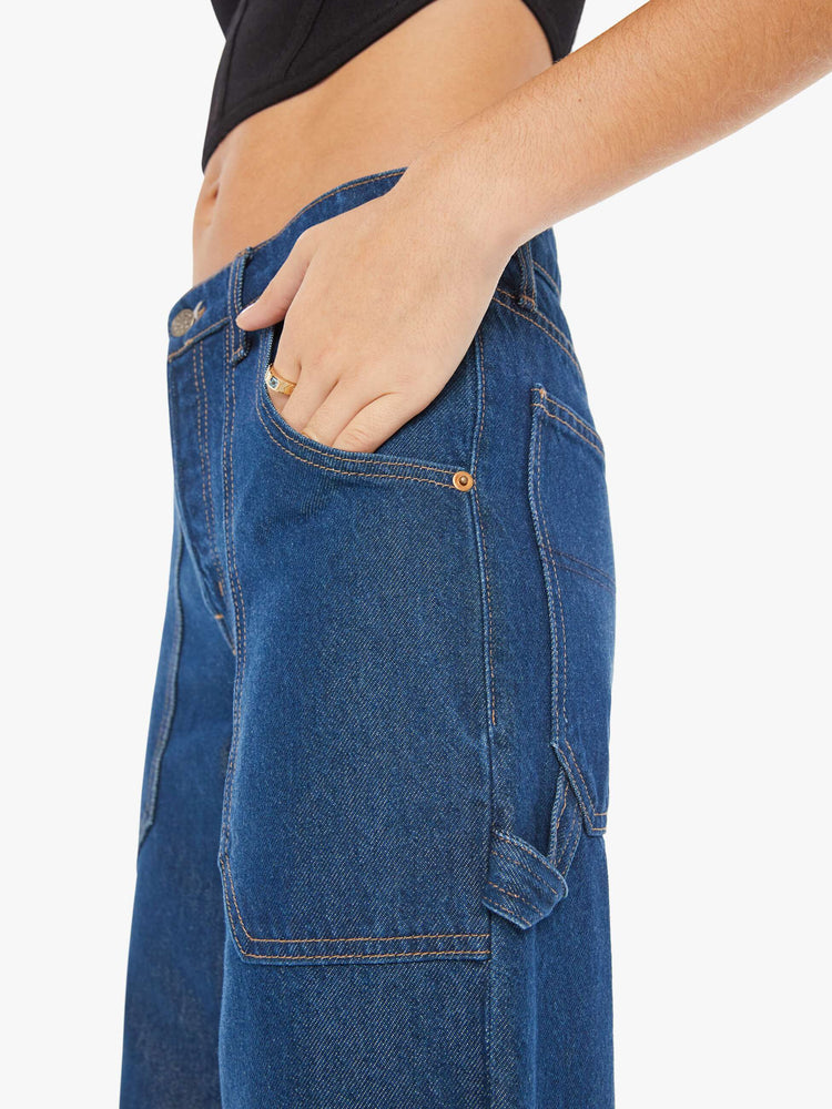 Side detail view of a womens dark blue denim jean featuring a long cuffed hem, a wide leg, carpenter details, and a loose slouchy fit.