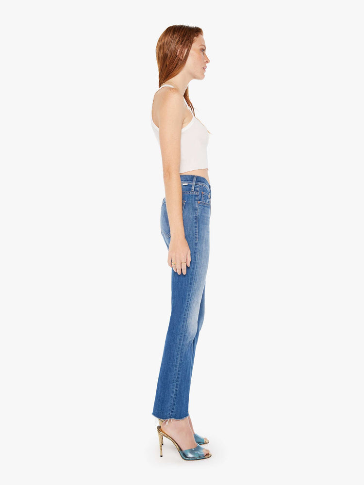 Side full body view of a woman wearing a medium blue wash jean featuring a high rise and a flood length raw bootcut hem.