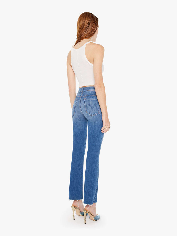 Back full body view of a woman wearing a medium blue wash jean featuring a high rise and a flood length raw bootcut hem.