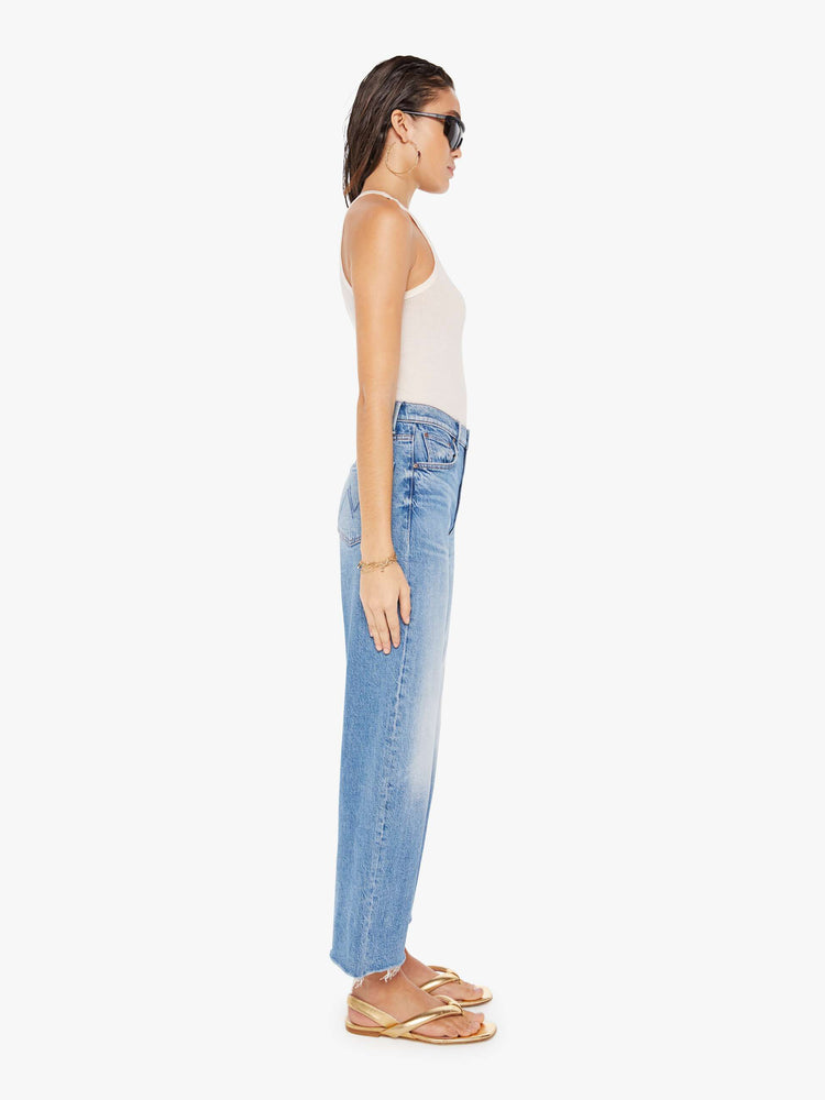 Side full body view of a woman wearing a medium blue wash jean featuring a high rise and an angle length wide leg with frayed hem.