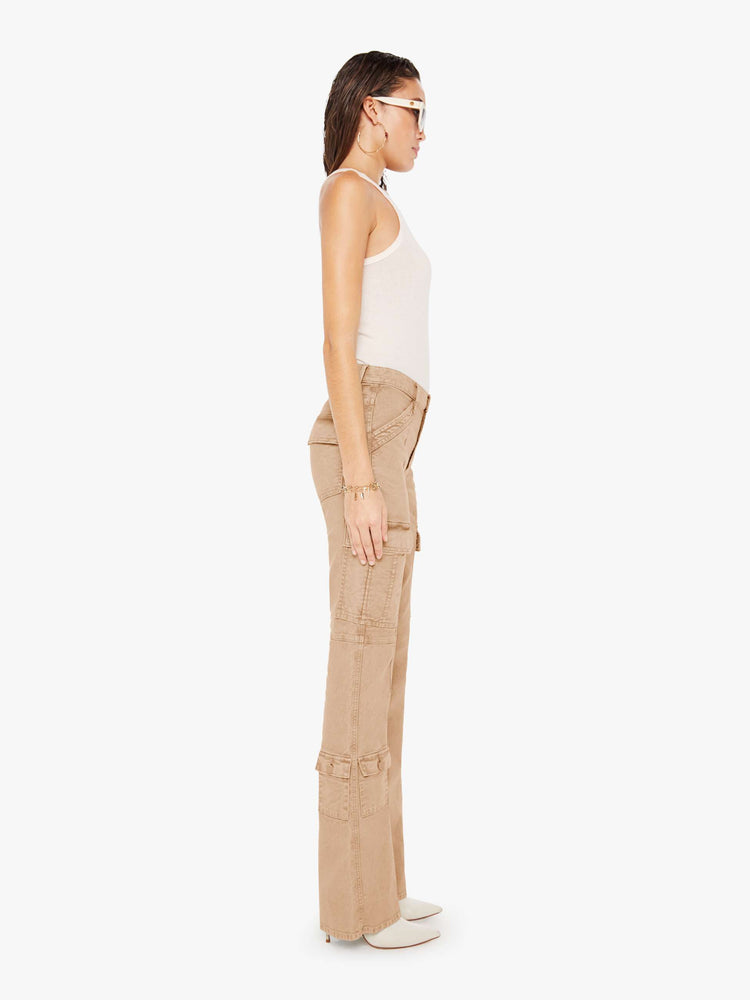 Side view of a woman wearing a light brown wash pant featuring darker brown hues around the seams, a high rise, a full length flare, and cargo pockets, paired with a white tank top.