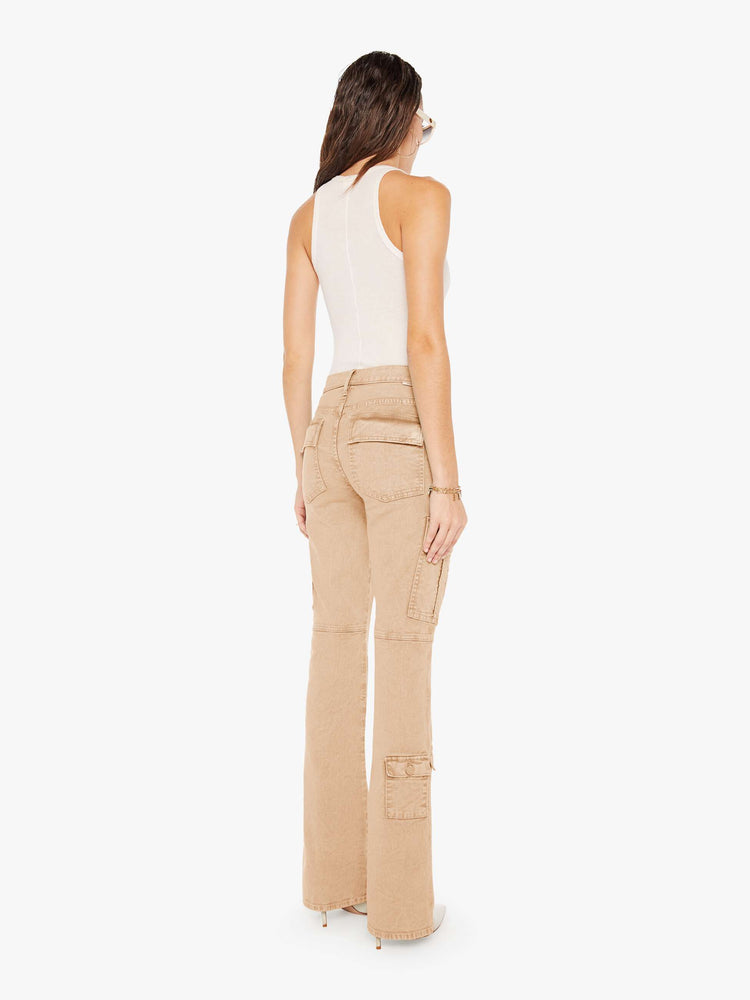 Back view of a woman wearing a light brown wash pant featuring darker brown hues around the seams, a high rise, a full length flare, and cargo pockets, paired with a white tank top.