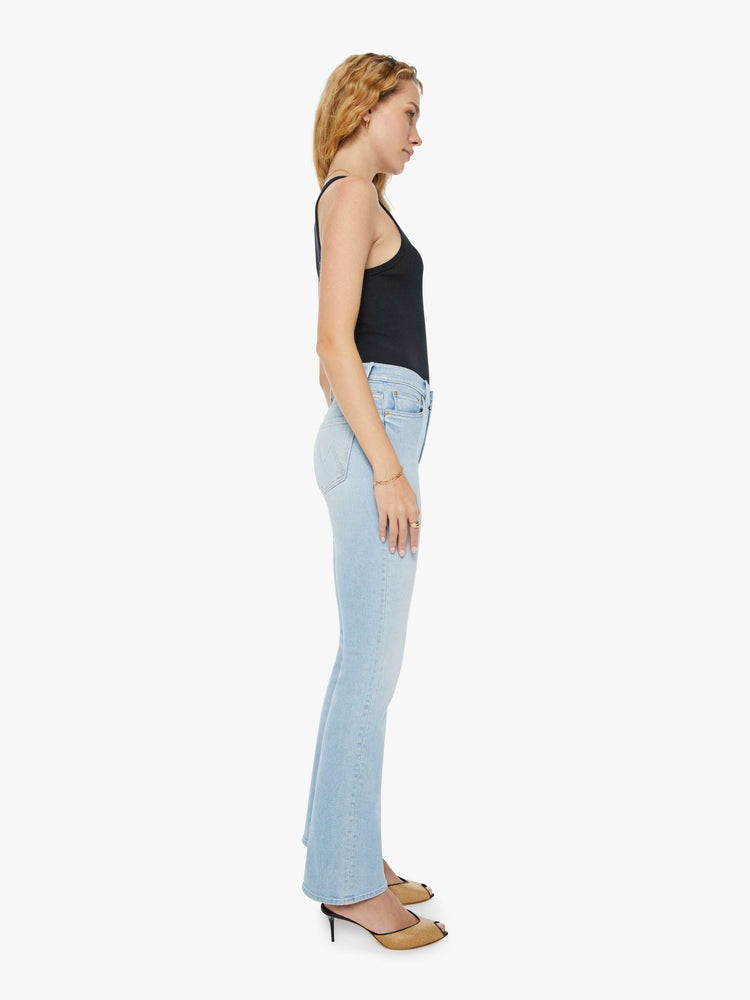 Side view of a woman in a high-rise light blue bootcut jean with whiskering and fading. Paired with a black tank top and mules.