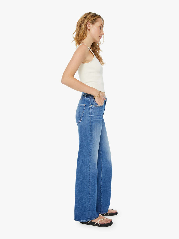 Side view of a woman in mid-blue wide leg jeans with whiskering and fading at the knees. Styled with a white tank top.