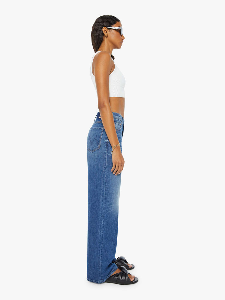 Side view of a woman in dark blue slouchy, high-waisted jeans with a loose wide leg and whiskering and fading at the knee. Styled with a white tank top.