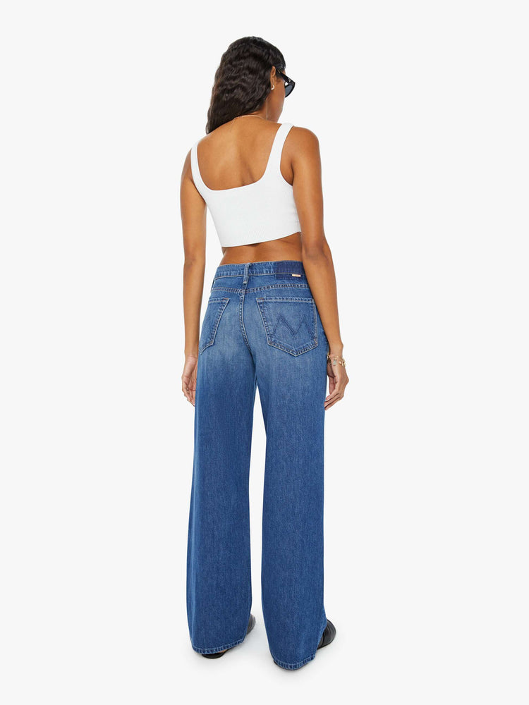 Back view of a woman in dark blue slouchy, high-waisted jeans with a loose wide leg and whiskering and fading at the knee. Styled with a white tank top.