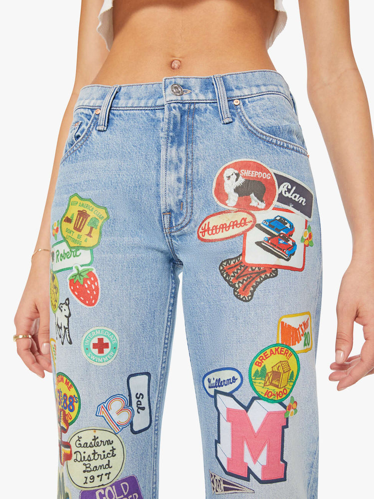 Front close up view of a woman in a light blue and colorful vinyl patches thoughout high-rise bootcut jean with a zip fly, looser leg and a 32-inch inseam with a clean hem. 