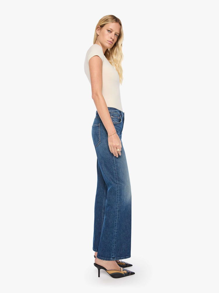 Side view of a woman in mid-blue mid-rise bootcut with a zip fly, looser leg and a long 32-inch inseam with a clean hem.