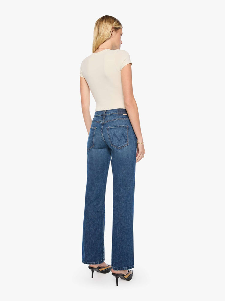 Back view of a woman in mid-blue mid-rise bootcut with a zip fly, looser leg and a long 32-inch inseam with a clean hem.