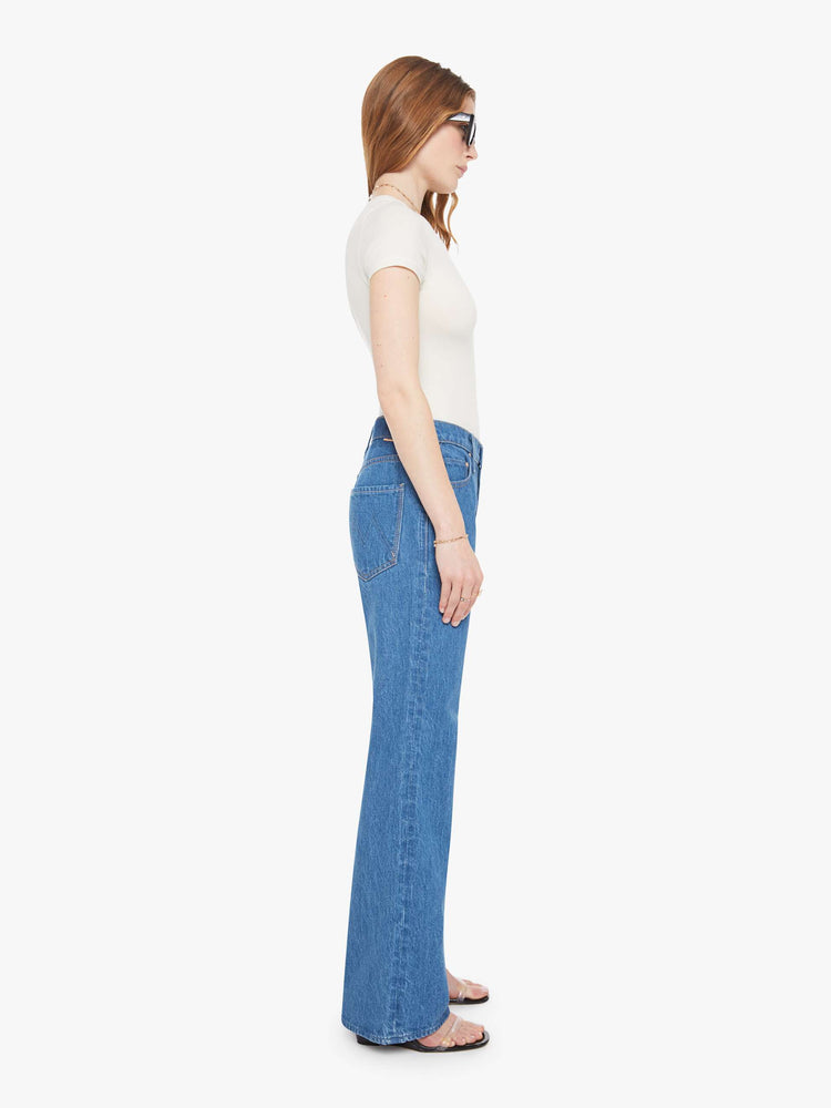 Side view of a woman in med blue high-rise bootcut with a zip fly, looser leg and a long 32-inch inseam with a clean hem.