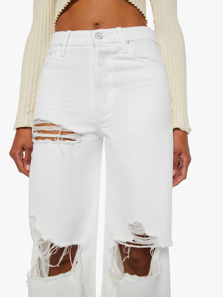 Detailed front view of a woman in off-white mid-rise jeans designed with a button fly, slouchy, loose fit  with a chewed hem, tears at the knees and thigh, finished with a pretzel-detailed button.