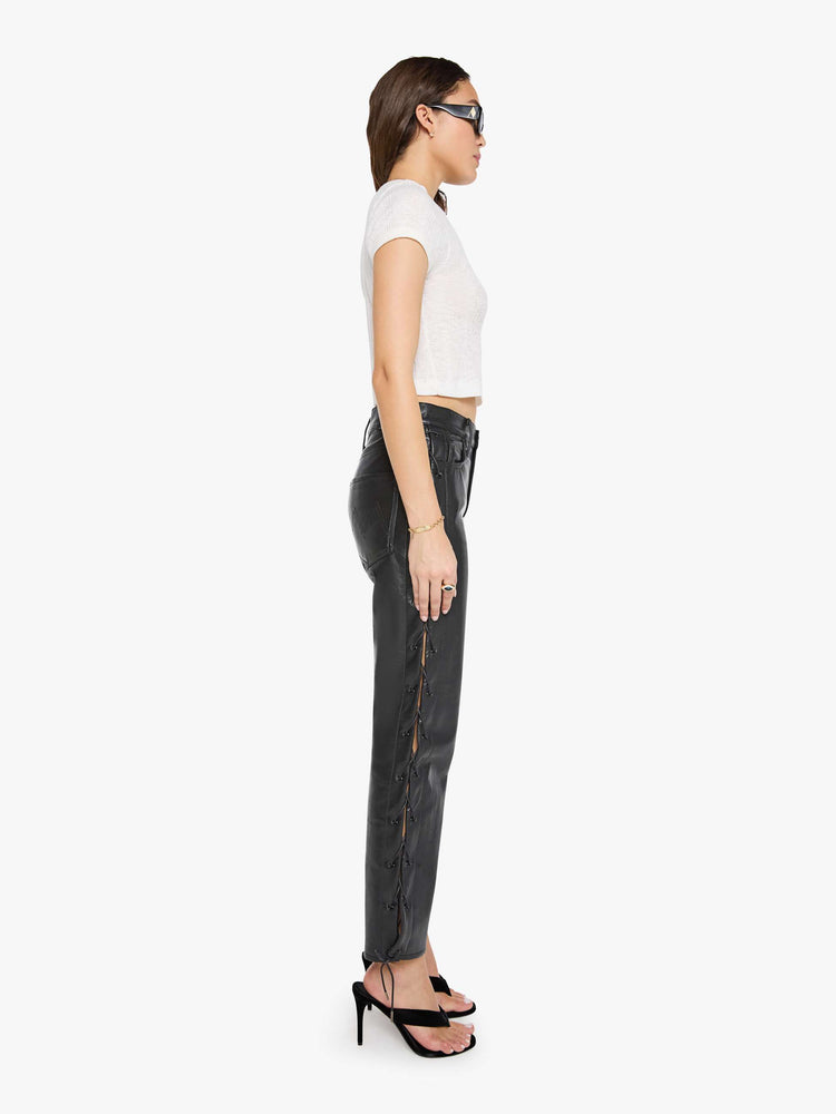 Side view of a woman in black leather high waisted pants with a zip fly, straight leg, 29-inch inseam and a lace-up detail on the outer seams.