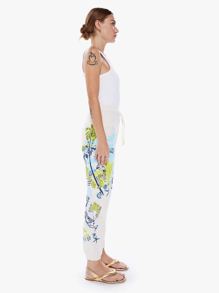 Side view of a woman high-rise drawstring waist and an ankle-length inseam with elastic hems sweatpants in off-white hue with a blue and green graphic.