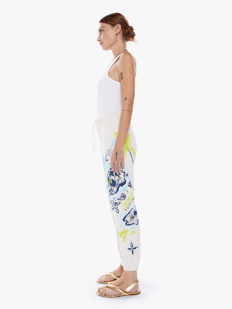 Left side view of a woman high-rise drawstring waist and an ankle-length inseam with elastic hems sweatpants in off-white hue with a blue and green graphic.
