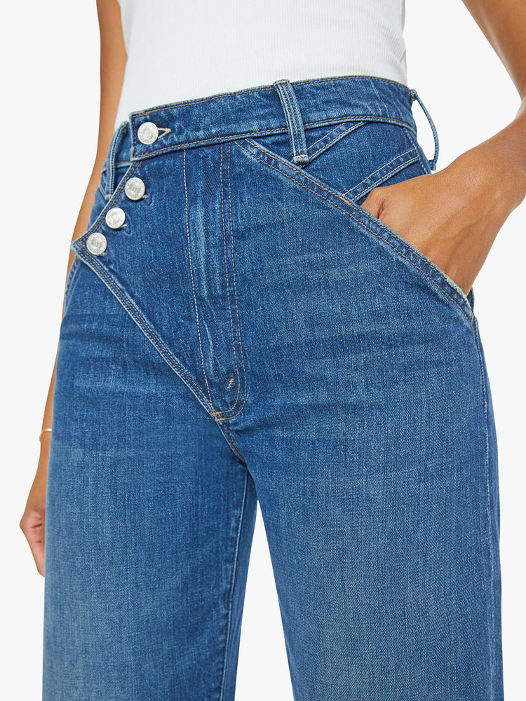 Waist close up view of a woman in a mid blue super high-rise straight-leg jeans with a buttoned flap at the waistband, angled seamed details and a 29-inch inseam with a clean hem.