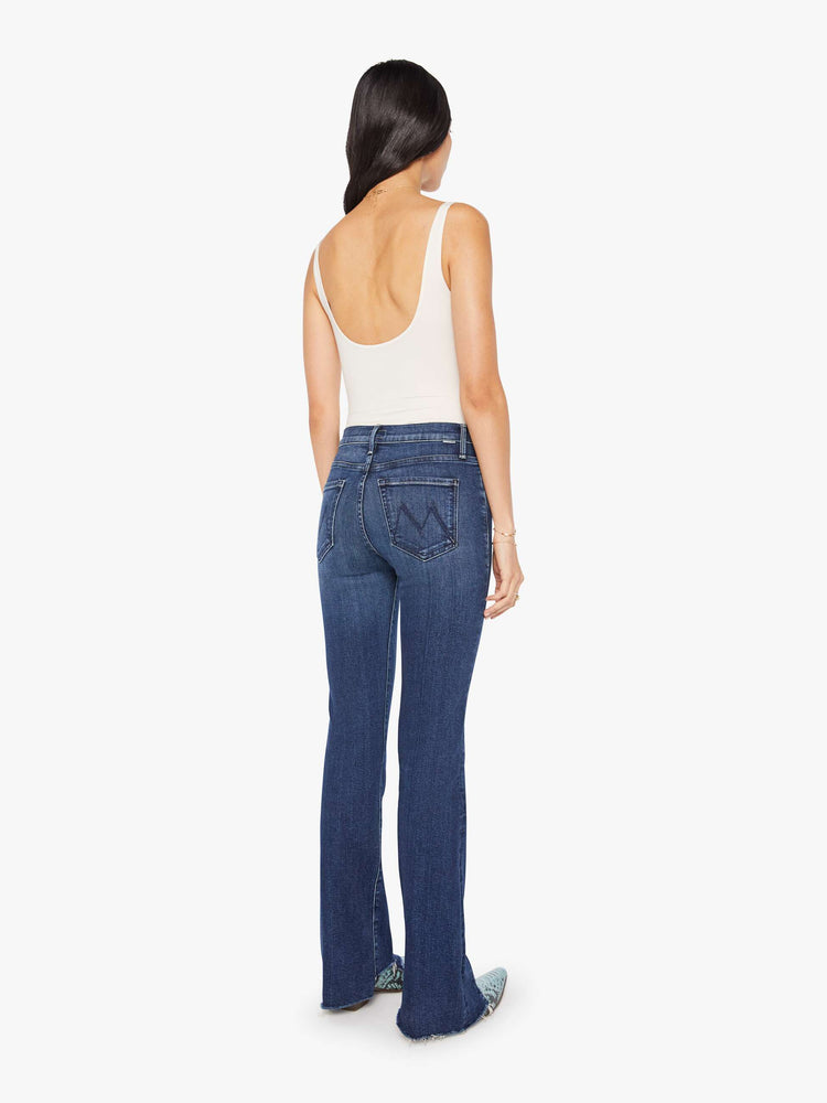 Back view of a woman in a dark blue high-waisted bootcut features a zip fly, seamed details down the front, a 31-inch inseam and a frayed triple step-hem.
