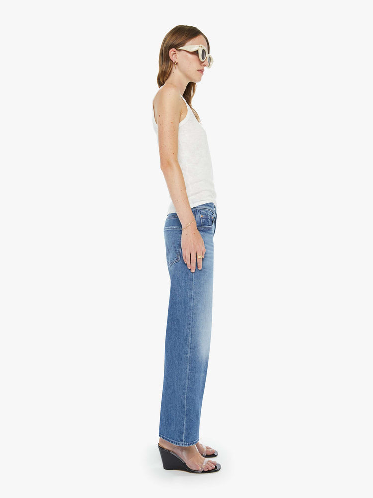 Side view of a woman in a mid blue mid-rise jeans with a loose fit, wide leg, zip fly and ankle-length inseam with a clean hem.
