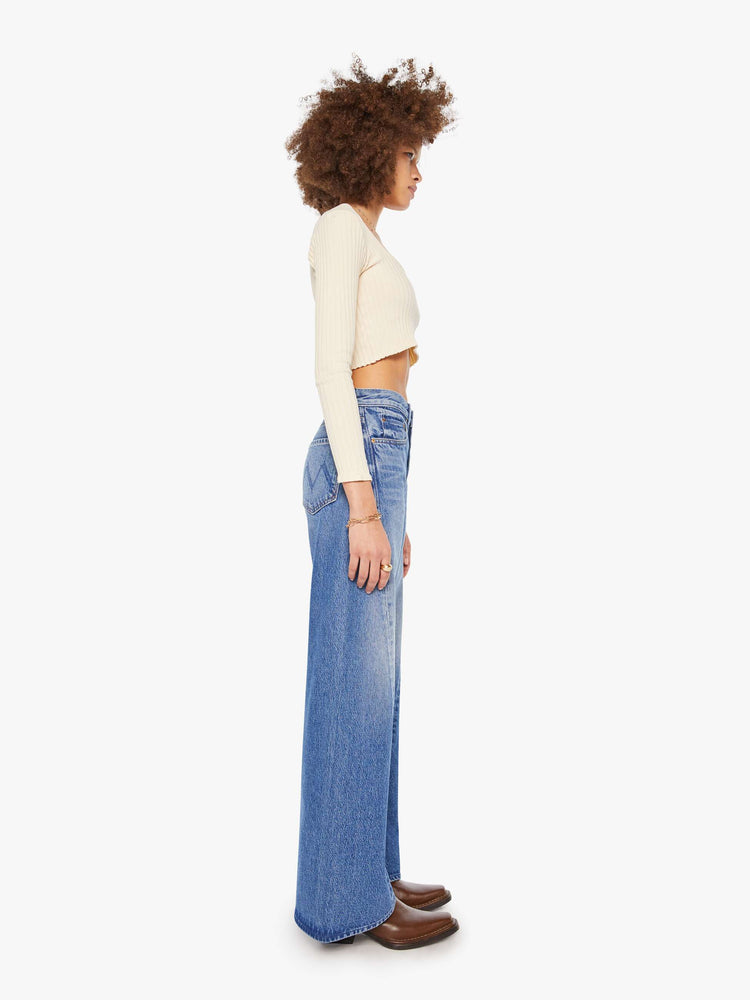 Side view of  woman in a mid blue high waisted jean with a wide leg  with a curved outer seam and a long 33.5-inch inseam with a clean hem. 