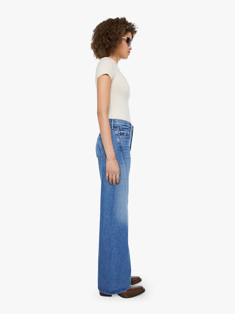 Side view of  woman in a mid blue high waisted jean with a wide leg  with a curved outer seam and a long 33.5-inch inseam with a clean hem. 