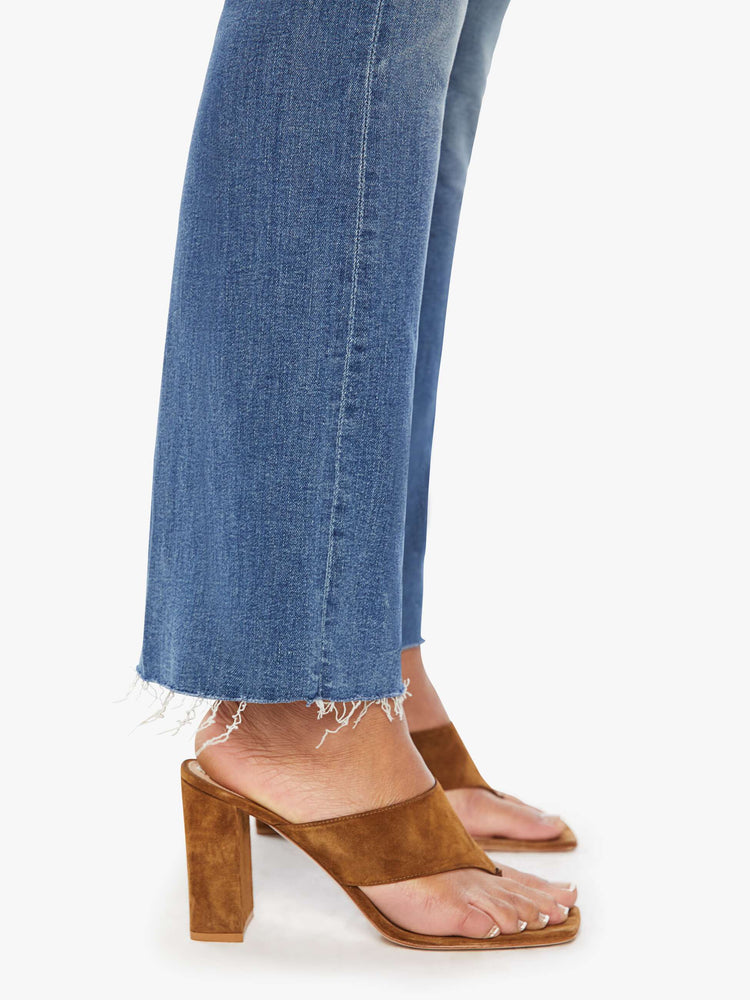 Side close up view of a womens medium blue wash jean featuring a high rise, flare leg, and a raw ankle length hem.