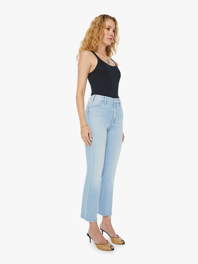 Angled view of a woman in light-blue high-rise flare that has an ankle-length inseam and a raw hem with subtle whiskering and fading throughout. Paired with a black tank top and heeled mules.