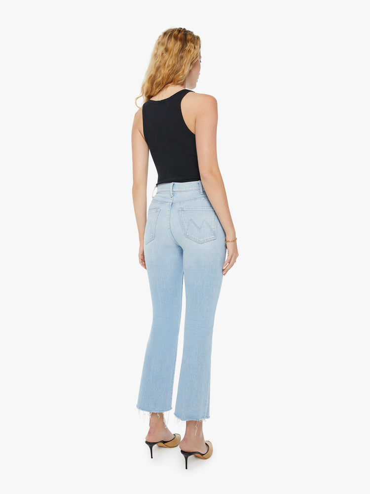 Back view of a woman in light-blue high-rise flare that has an ankle-length inseam and a raw hem with subtle whiskering and fading throughout. Paired with a black tank top and heeled mules.