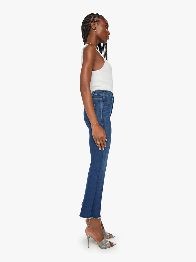 Side view of a womens dark blue wash jean featuring a high rise and an ankle length flare with raw hem.