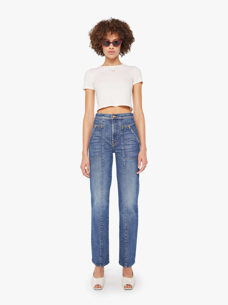 Front view of a woman in a mid-blue high-waisted jeans with a wide straight leg, zip fly, patch pockets with rivet details, a seam down the front and back and a clean 32-inch inseam.