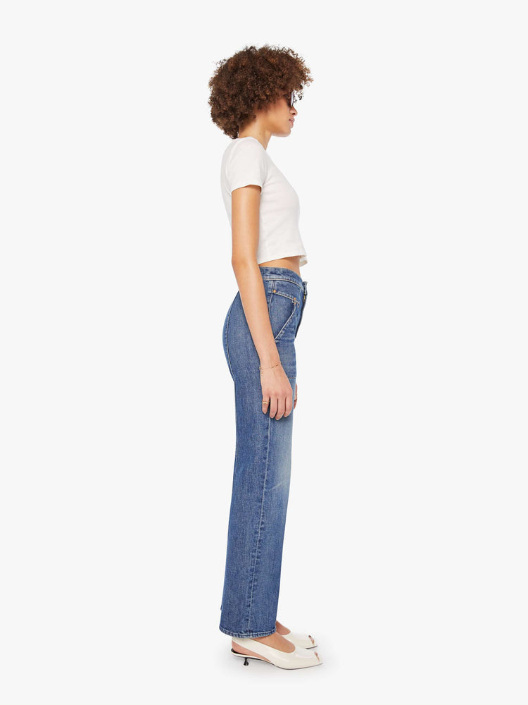 Side view of a woman in a mid-blue high-waisted jeans with a wide straight leg, zip fly, patch pockets with rivet details, a seam down the front and back and a clean 32-inch inseam.