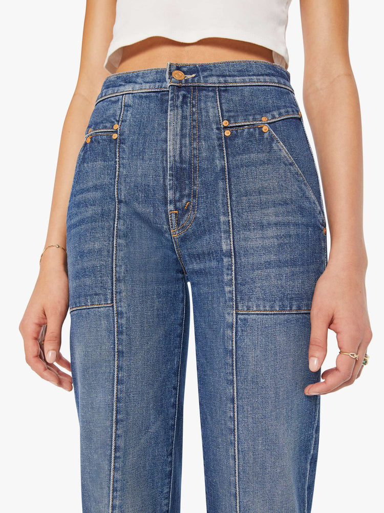 Close up view of a woman in a mid-blue high-waisted jeans with a wide straight leg, zip fly, patch pockets with rivet details, a seam down the front and back and a clean 32-inch inseam.
