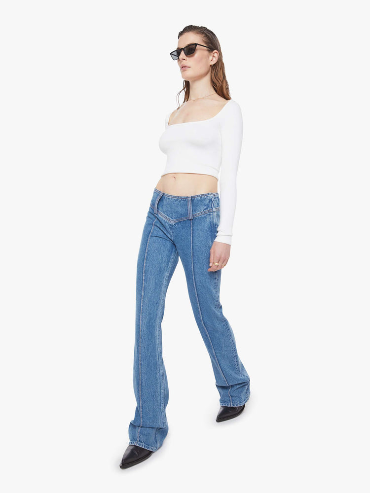 Walking view of a woman in a mid blue  low-rise jeans feature an extra-wide waistband with a side-zip closure, a slightly flared leg with pintuck details down the front and a long 34-inch inseam with a clean hem. 