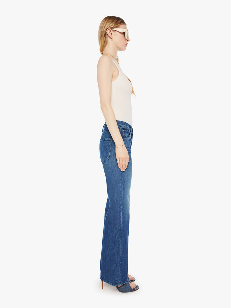 Side view of a woman in a med blue high-rise bootcut with a zip fly, looser leg and a long 31-inch inseam with a clean hem.