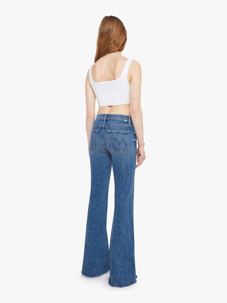 Back view of a woman in a medium blue high-rise flare with a zip fly, long 32-inch inseam and a frayed hem.