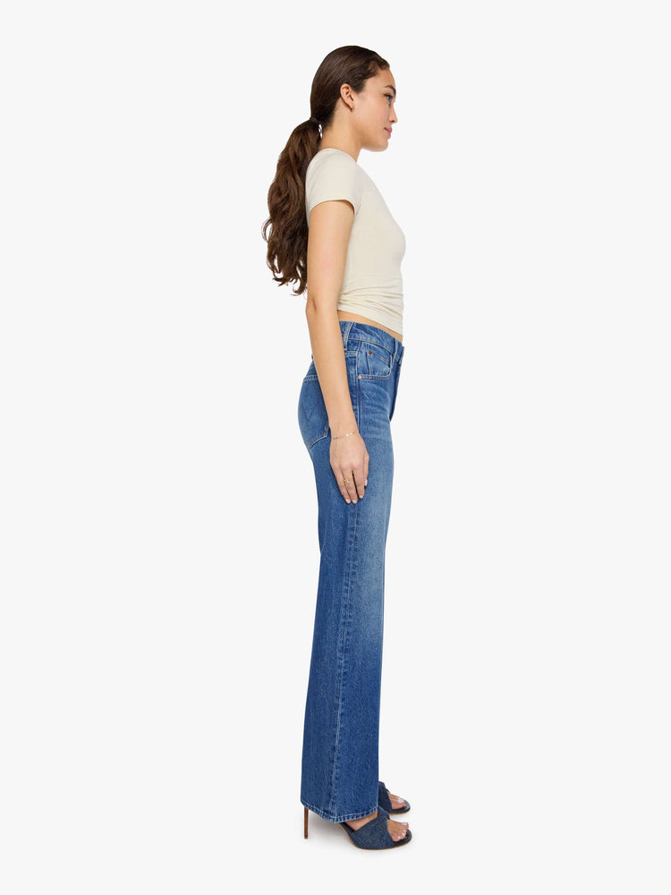 Side view of a woman in a med blue low-slung jeans designed with a high rise, zip fly, a 32-inch inseam and a clean hem.
