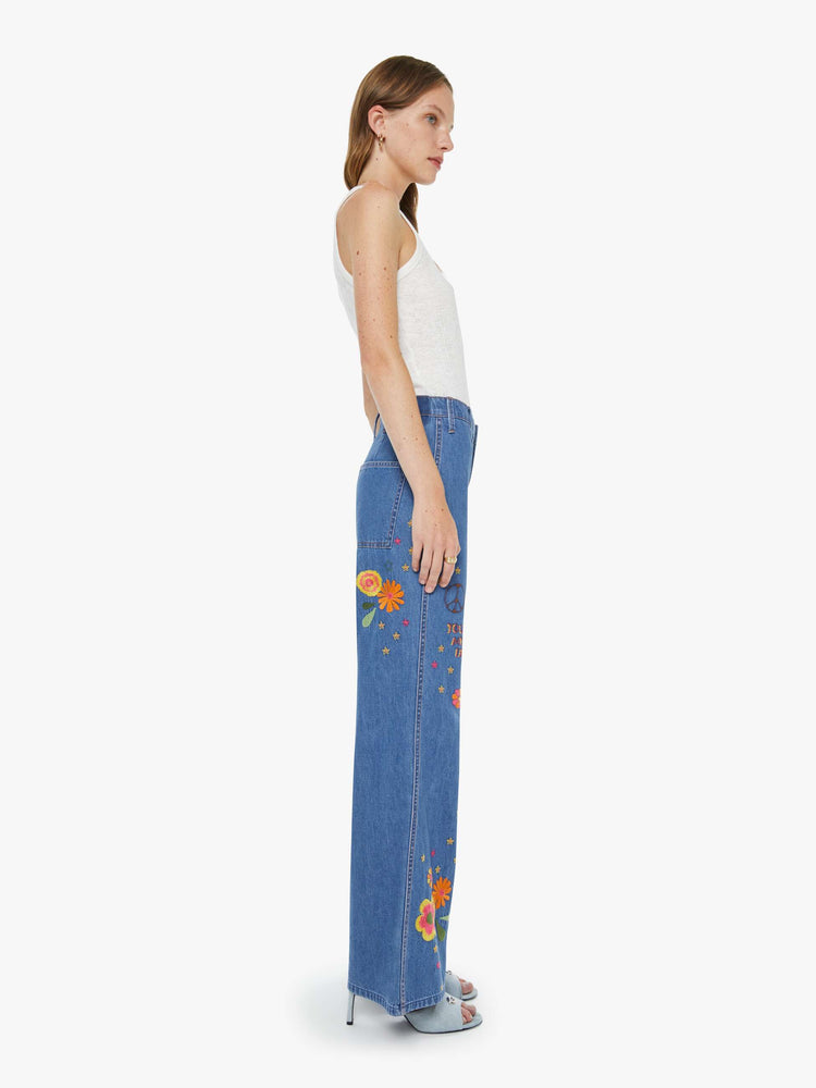 Side view of a woman in a med blue with embroidery flowers  wide leg jeans with a high rise, zip fly, long inseam and a clean hem.