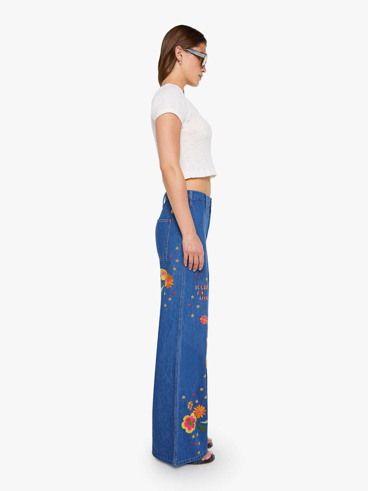 Side view of a woman in a med blue with embroidery flowers  wide leg jeans with a high rise, zip fly, long inseam and a clean hem.