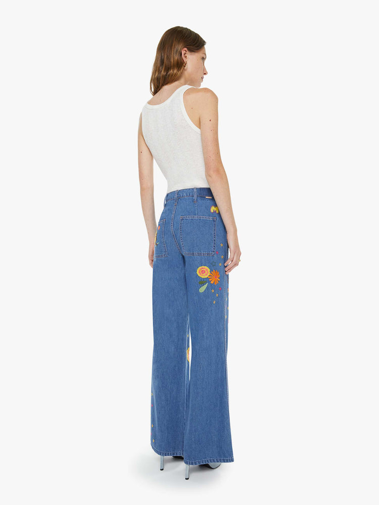 Back view of a woman in a med blue with embroidery flowers  wide leg jeans with a high rise, zip fly, long inseam and a clean hem.
