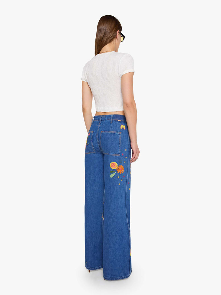 Back view of a woman in a med blue with embroidery flowers  wide leg jeans with a high rise, zip fly, long inseam and a clean hem.