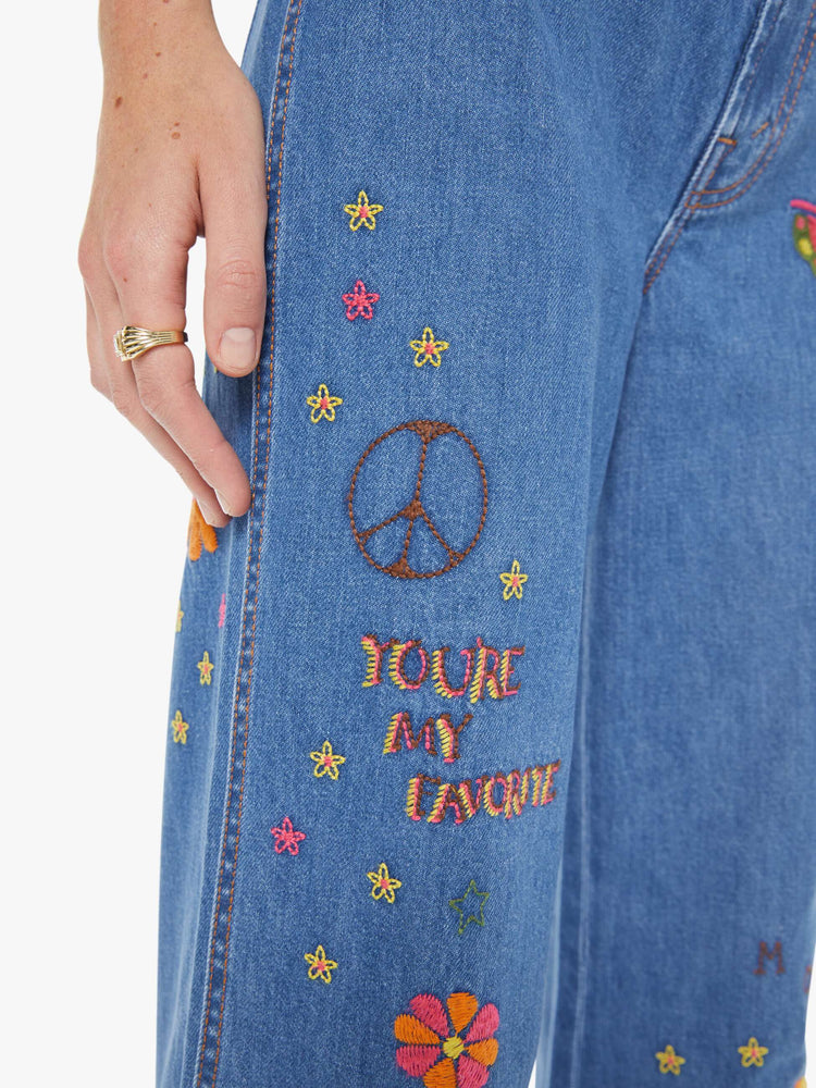 Close up view of a woman in a med blue with embroidery flowers  wide leg jeans with a high rise, zip fly, long inseam and a clean hem.